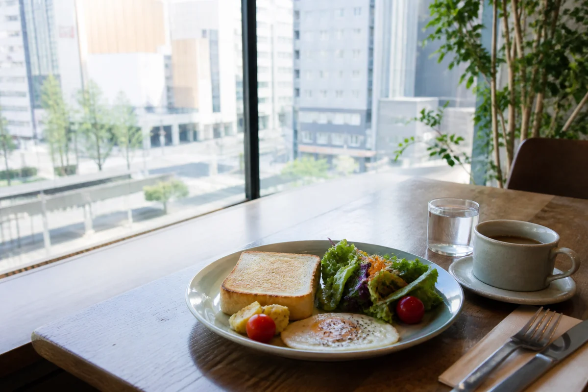 都市の景色を望む明るいカフェで、トーストや卵、サラダを盛り合わせた朝食のイメージ